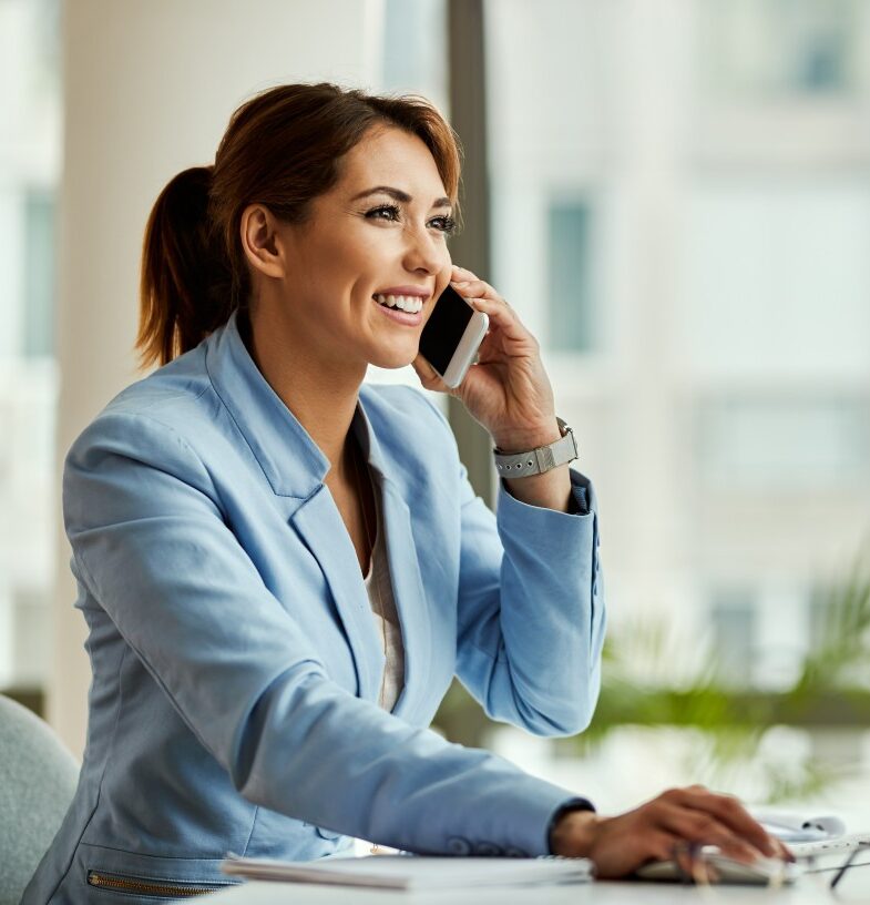 jovem_empresaria_feliz_trabalhando_no_pc_desktop_e_se_comunicando_por_telefone_celular_no_escritorio-24453459